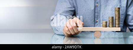 Close-up di un imprenditore la mano di bilanciamento delle monete impilate su altalena in legno con il dito sulla scrivania Foto Stock