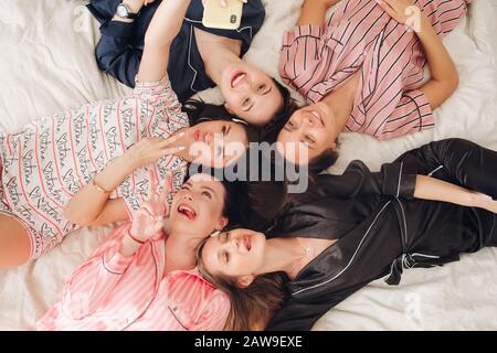 Vista dall'alto del gruppo di donne piuttosto giovani in pigiami sdraiati a letto e divertirsi. Concetto di festa di compleanno Foto Stock