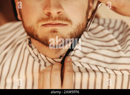 Parte inferiore del viso barbuto uomo in pelignoir a righe uomini barbiere primo piano, barba forbice Foto Stock