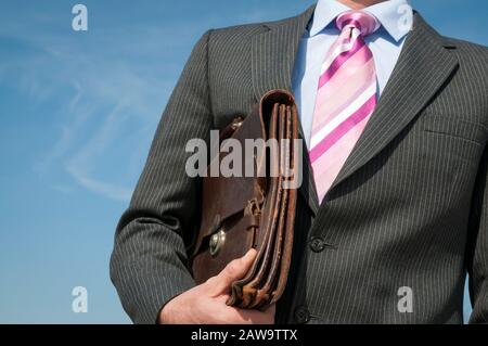 Uomo d'affari irriconoscibile che si trova all'aperto in abito grigio con cravatta rosa che tiene la valigetta davanti a un cielo blu brillante Foto Stock