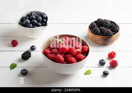 tre ciotole con frutti di bosco, lampone, mirtilli, more, su sfondo di legno bianco Foto Stock