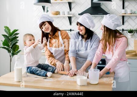 Donna sorridente di mezza età in grembiule da cucina che arrotola l'impasto e due figlie che la aiutano. Figliola baby ragazza seduta sul tavolo e divertirsi. Felice Foto Stock