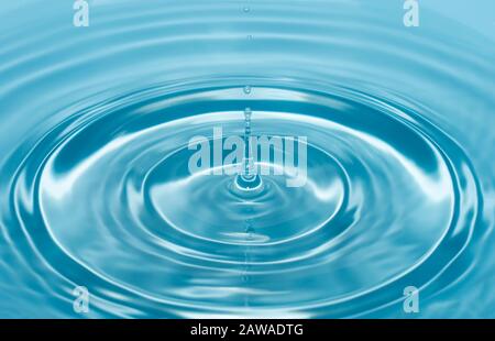 una goccia d'acqua cade con uno spruzzo in acqua chiara e blu che forma cerchi sull'acqua. Foto Stock