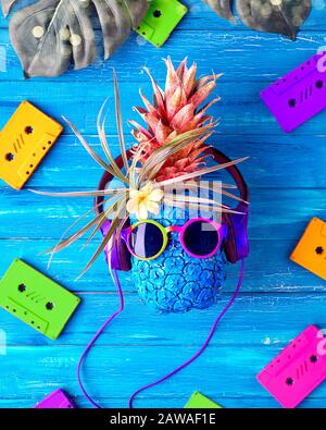 Concept di musica da discoteca retro 90s. Il carattere dell'ananas dell'ippopotami negli occhiali da sole e negli auricolari rossi ascolta la musica. Legno rustico blu con audio neon 90s Foto Stock