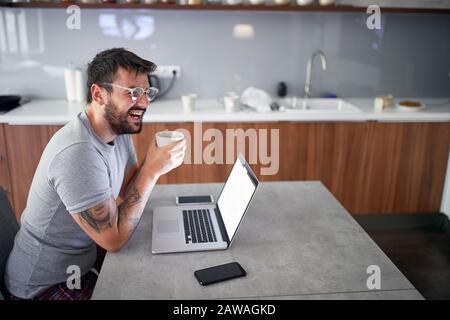giovane uomo adulto con occhiali da barba, sorridente davanti al portatile sul tavolo, con una tazza. moderno, casual, stile di vita, concetto di business Foto Stock