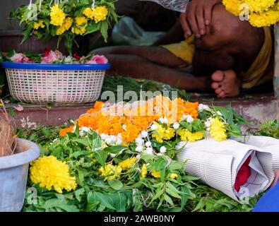 Fiori arancioni, rossi e gialli circondati da foglie verdi, giacenti a terra accanto al venditore seduto vicino all'ingresso di un tempio. Foto Stock