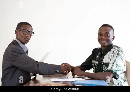 Uomini d'affari africani scuotendo le mani in ufficio sigillando un affare ridendo e sorridendo con le camicie e le tute tradizionali a Bamako, Mali Foto Stock
