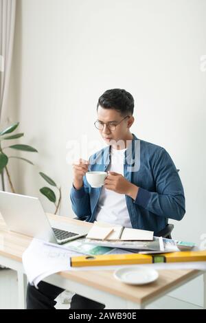 Bel giovane che beve caffè e che lavora alla sua scrivania in ufficio Foto Stock