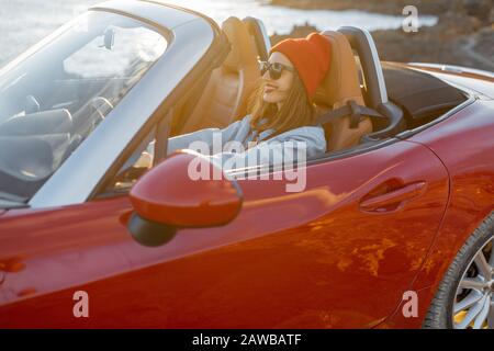 Stile di vita ritratto di donna felice in cappello rosso guida auto convertibile sulla costa rocciosa dell'oceano durante un tramonto. Stile di vita spensierato e concetto di viaggio in auto Foto Stock