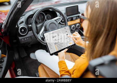 Giovane donna che controlla l'auto utilizzando un tablet digitale e un'applicazione mobile mentre si siede sul sedile del conducente, vista dal retro Foto Stock