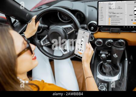 Giovane donna che controlla l'auto utilizzando lo smartphone con applicazione mobile lanciata, seduto sul sedile del conducente, vista dal retro Foto Stock