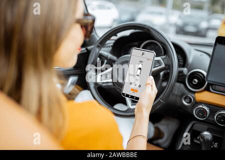Giovane donna che controlla l'auto utilizzando lo smartphone con applicazione mobile lanciata, seduto sul sedile del conducente, vista dal retro Foto Stock
