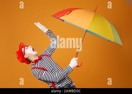 mimo mad uomo triste che guarda in su con ombrello multi-colorato in mano isolato su sfondo arancione vuoto in studio Foto Stock
