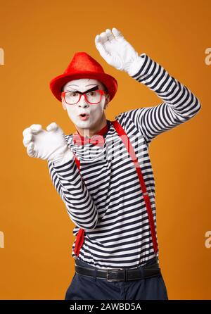 Giovane uomo mime in guanti bianchi e cappello rosso che guarda la fotocamera su sfondo arancione vuoto Foto Stock