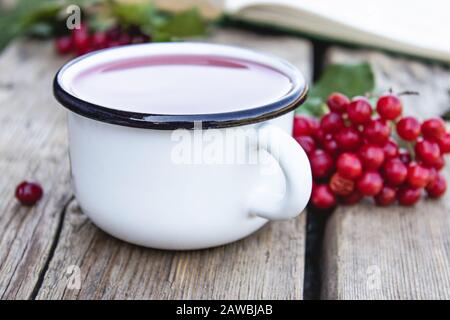 Tazza bianca o tazza di tè vituro caldo su un tavolo di legno vicino a un libro aperto e frutti rossi di vituro. Fonte di vitamine naturali. Usato nella medicina popolare. Foto Stock