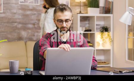 Bell'imprenditore prendere un sorso di caffè mentre si lavora su un computer portatile in soggiorno. La ragazza in background parla al telefono. Foto Stock