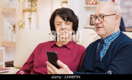 Felice coppia senior con una videochiamata seduto sul divano nel soggiorno. Coppie di età che utilizzano la tecnologia moderna Foto Stock