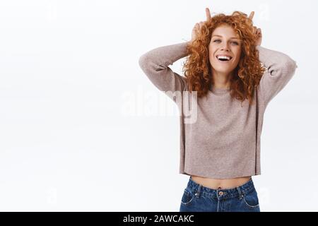 Concetto di benessere, emozioni e stile di vita. Ritratto attraente sassoso redhead curly donna in blouse trendy, mostrando corna, tenere indice dita dietro Foto Stock