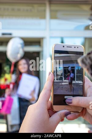 Punto di vista di una persona che scatta una foto con uno smartphone di una ragazza che porta fiori e un diploma. Celebrazione di laurea, donne sucessfull in busine Foto Stock