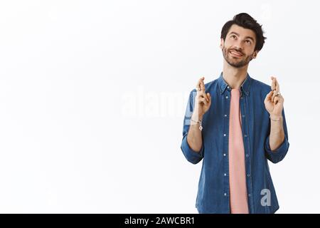 Sognante affascinante ragazzo 25s con bearded in camicia hippster, t-shirt guardando daydreaming o fantasticing, cross fingers per la buona fortuna, facendo desiderio, sorridendo Foto Stock