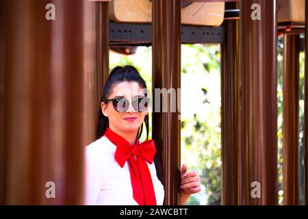 Bruna attraente in un vestito scuola ragazza che si posa in un parco pubblico circondato da tubi di rame marrone. Grande sciarpa rossa su una camicia bianca Foto Stock