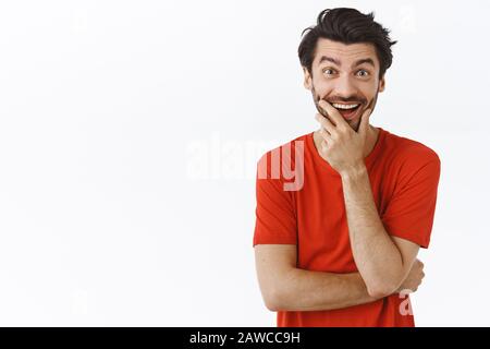 Girovita ritratto gioioso entusiasta giovane bearded uomo in t-shirt rossa, ridere e toccare setole come sempre curioso, stare intrattenuto e divertito Foto Stock