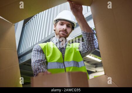 Giovane lavoratore manuale nella confezione del casco da lavoro o disimballaggio della scatola di cartone in magazzino Foto Stock