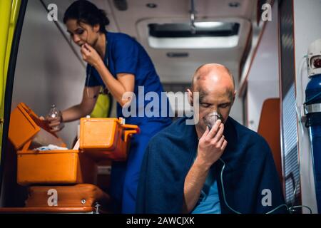 L'uomo respira attraverso la maschera di ossigeno, giovane infermiera in uniforme medica apre qualcosa preso dalla borsa medica nell'automobile dell'ambulanza Foto Stock