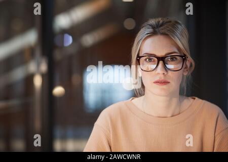 Ritratto di giovane donna con capelli biondi e occhiali che guarda la macchina fotografica mentre si siede in ufficio Foto Stock
