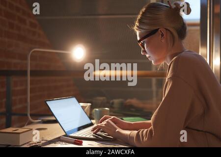 Giovane donna bionda in occhiali e in abbigliamento casual seduto alla scrivania e lavorando su laptop fino a tardi in ufficio Foto Stock