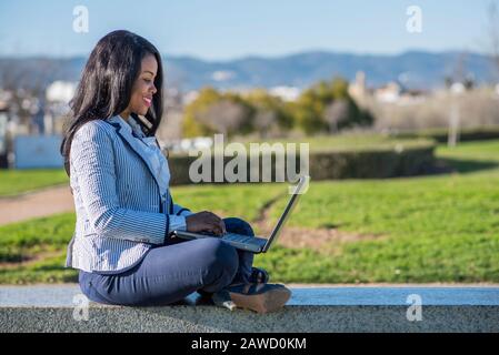 Donna afro-americana seduta su una panchina utilizzando un computer portatile in un parco all'aperto Foto Stock