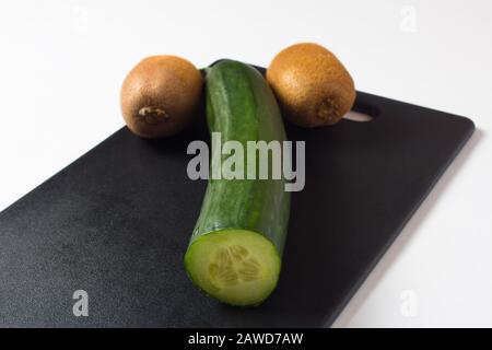 Un cetriolo e due kiwi si trovano su un tagliere nero. Alimenti dietetici e insalate per uno stile di vita sano. La somiglianza di un simbolo fallico o scava maschile Foto Stock