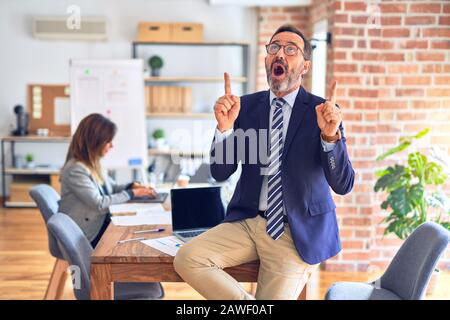 Bell'uomo d'affari di media età che indossa gli occhiali seduti sulla scrivania in ufficio stupito e sorpreso guardando in alto e indicando con le dita e braccio sollevato Foto Stock