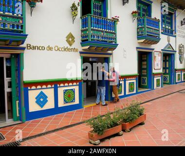 Uomini che si trovano al di fuori di una banca in stile coloniale e edificio governativo decorato con zocalos (fregi) sulla piazza principale di Guatape, Antiquia, Colombia Foto Stock