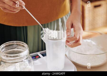 Immagine ravvicinata della donna che mette il cucchiaio di lye in vaso di plastica quando fa il sapone a casa Foto Stock
