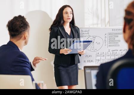 Una donna seria che legge il suo rapporto annuale ai colleghi riuniti nella sala riunioni Foto Stock