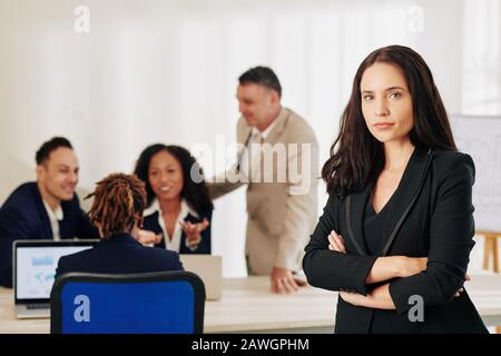Bella imprenditore femminile di successo in piedi con le braccia piegate, il suo team di discutere di lavoro in background Foto Stock