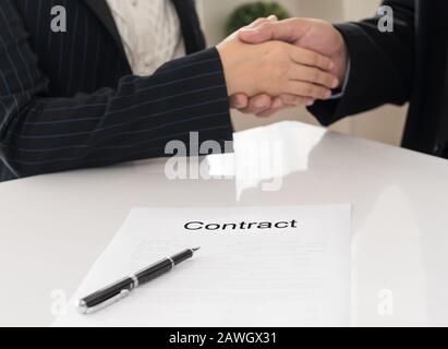 Documento contrattuale di primo piano con immagine di una stretta di mano tra due partner commerciali che firmano un contratto in background. Foto Stock