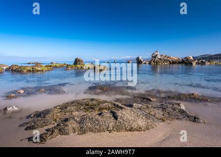 Rocce lungo la spiaggia e un tranquillo mare mediterraneo vicino l'Ile Rousse nella regione Balagne della Corsica in una giornata di sole con il deserto des Agriat Foto Stock