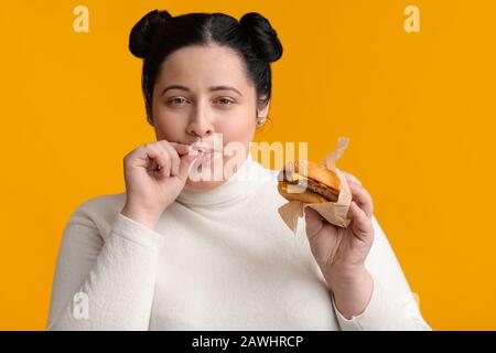 Giovane obesa ragazza mangiare hamburger e leccare le dita, godendo fast food Foto Stock