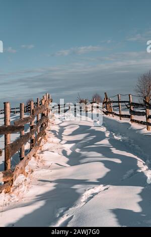Stagione invernale, immagine paesaggistica di una strada di villaggio coperta da neve al tramonto, paesaggio di campagna con recinzione in legno, cielo nuvoloso, alberi congelati e footpa Foto Stock
