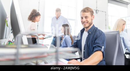 Giovane uomo ridendo, seduto ad una scrivania in ufficio e lavorando su un nuovo business. Foto Stock