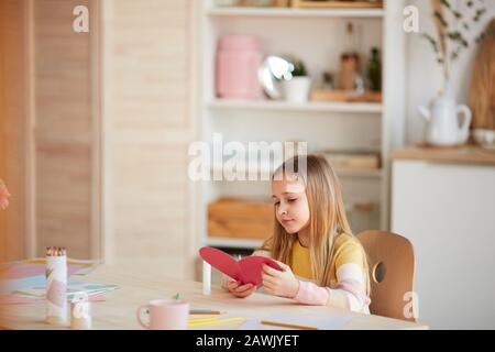 Ritratto caldo-tonato di cute ragazza che fa la scheda a forma di cuore e sorridente mentre seduto al tavolo in casa accogliente interno, copia spazio Foto Stock
