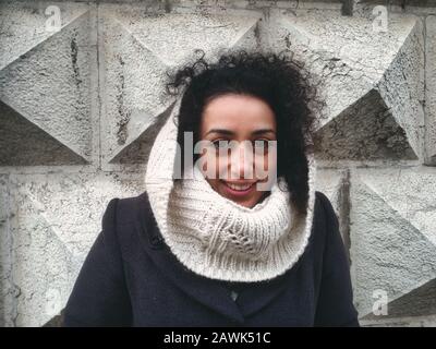 Bella giovane donna caucasica con lunghi capelli ricci neri in abiti invernali in piedi contro un muro di pietra edificio Foto Stock