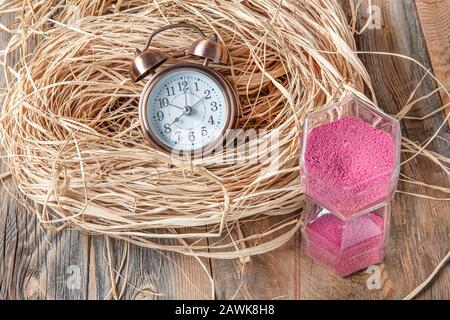 Clessidra rosa e sveglia su paglia su pavimento in legno. Concetto Di Tempo Foto Stock