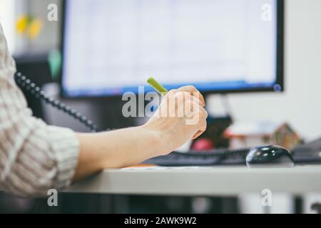 Primo piano della mano di una donna con una penna sullo sfondo del monitor. È al telefono. Giorni lavorativi in ufficio. Macro di messa a fuoco selettiva con poco profonda Foto Stock
