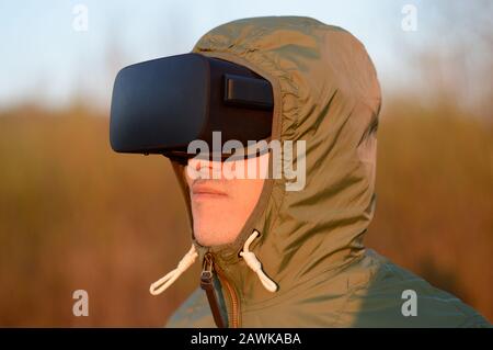 Ritratto di un giovane felice con cuffia per realtà virtuale e cappuccio protettivo verde all'aperto al tramonto Foto Stock