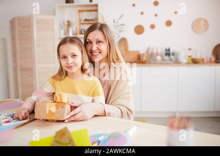 Ritratto caldo-tonato di madre felice che posa con la figlia mentre l'apertura presenta al tavolo della cucina di legno, spazio della copia Foto Stock