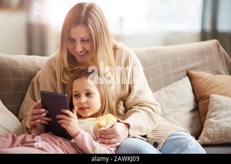 Ritratto caldo-toned di madre matura felice che gioca con ragazza cute mentre godendo il tempo insieme sul divano e usando il tablet digitale, spazio della copia Foto Stock