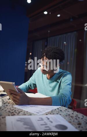 Ritratto della vista laterale del giovane uomo d'affari africano che lavora in ritardo mentre si siede al tavolo in stanza oscura e usando il tablet digitale, copia spazio Foto Stock